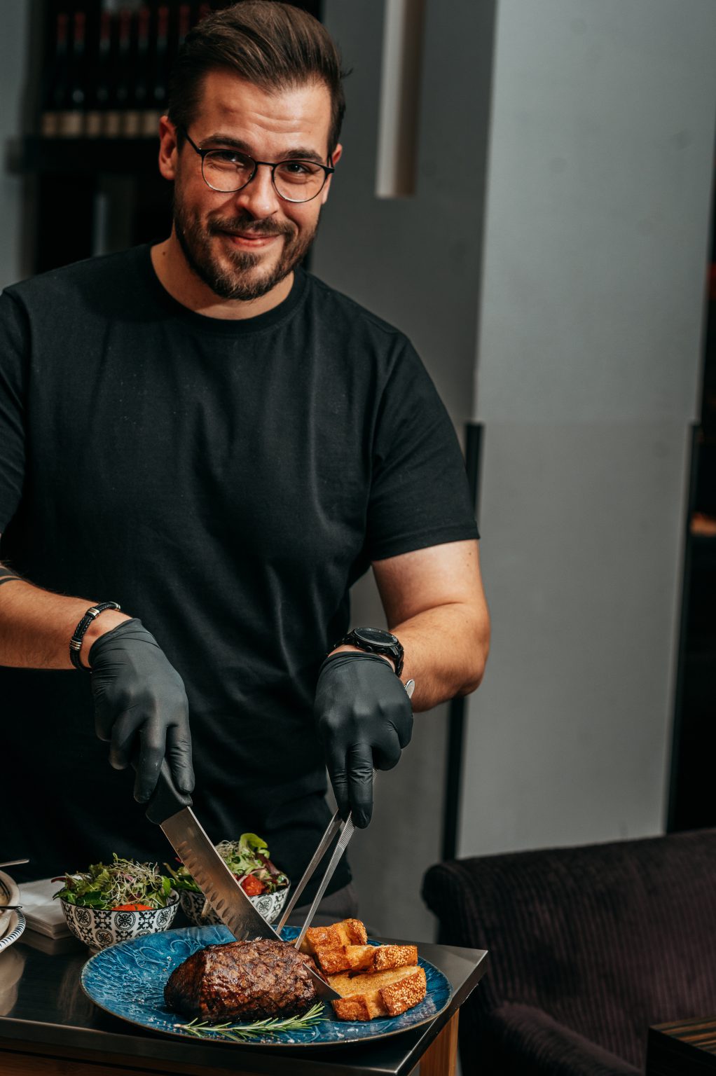 Ein Mann mit Brille und schwarzen Handschuhen serviert Chateaubriand am Tisch.