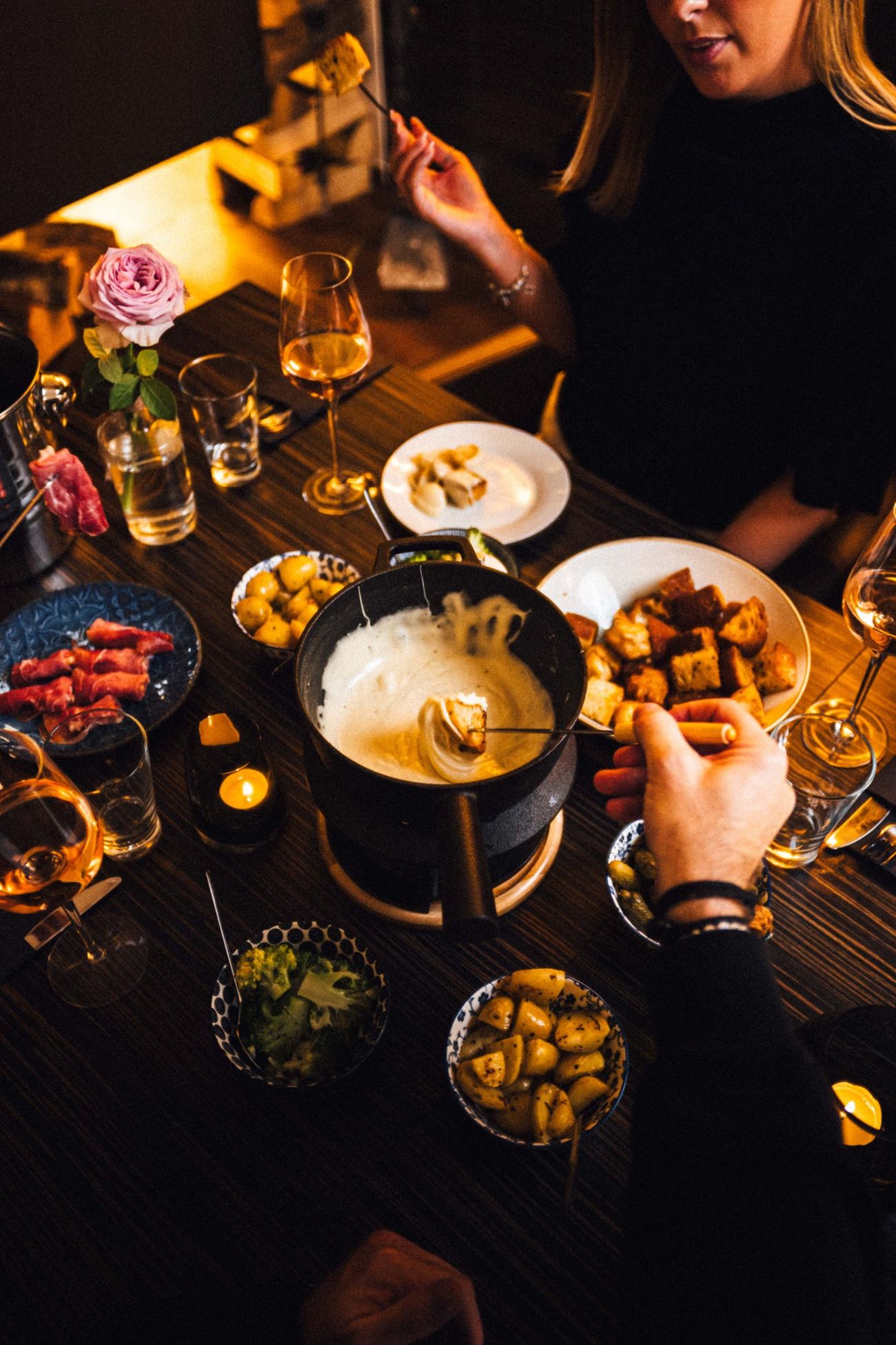 Gedeckter Tisch im Kerzenlicht mit Käsefondue, Landbrot, Kartoffeln, Brokkoli, Bündnerfleisch und Wein, eine Gabel taucht Brot in den Käse
