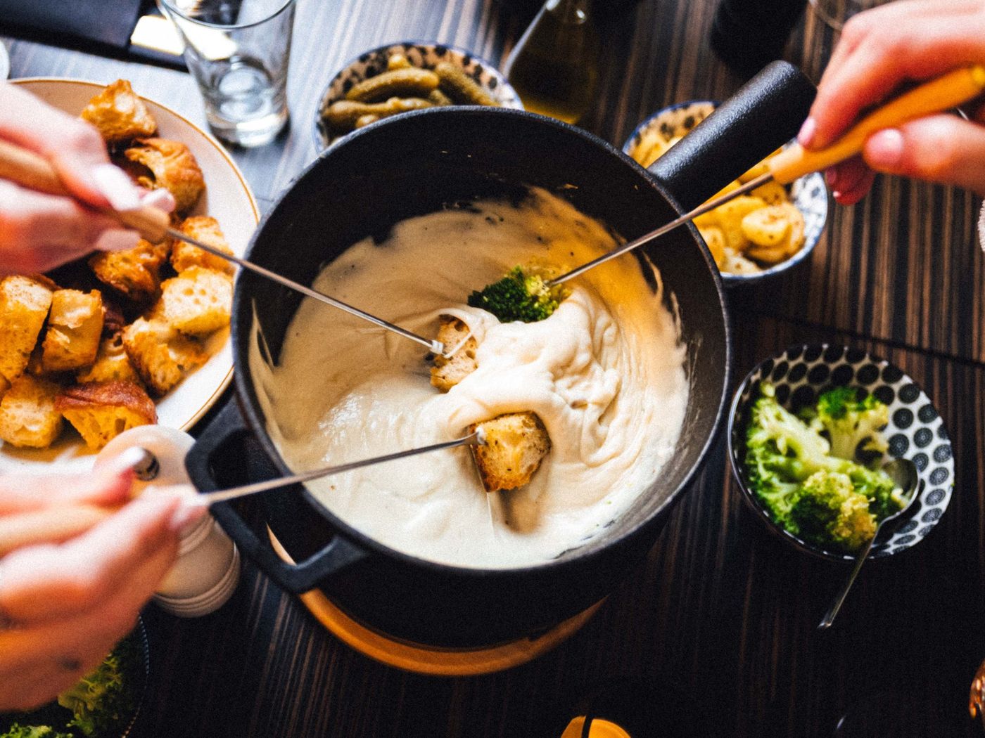 Fonduetopf von oben, rundherum Schalen mit Landbrot, Brokkoli, Kartoffeln und eingelegten Gurken